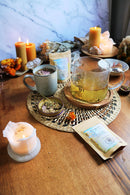 Eine Teekanne mit Kräutertee steht auf einem Tisch, umgeben von brennenden Kerzen, getrockneten Kräutern, einer Tasse und zwei Tüten Tee.