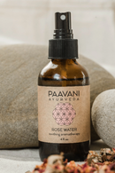 Eine braune Sprühflasche "PAAVANI AYURVEDA ROSE WATER soothing aromatherapy" (4 fl. oz.) steht zwischen großen grauen Steinen und verstreuten getrockneten Rosenblättern, was auf natürliche Inhaltsstoffe und Beruhigung hinweist.