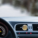 Ein silberfarbener Auto-Lufterfrischer mit einem Lebensbaum-Design und gelbem Hintergrund ist an der Lüftungsdüse eines Armaturenbretts befestigt. Die Ansicht ist vom Fahrersitz aus mit Blick auf eine verschneite oder neblige Straße.