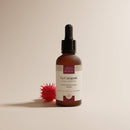 Eine gefrostete Glas-Tropfflasche mit „La Carapate“-Haaröl von Comptoir des Huiles neben einer roten, stacheligen Samenkapsel.