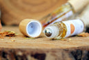 - Eine liegende Roll-on-Flasche mit Chakra-Öl und einem Metallrollerball liegt neben einer abgenommenen Holzkappe auf einer geschnittenen Holzoberfläche. Umher sind einige getrocknete Pflanzenreste verstreut.