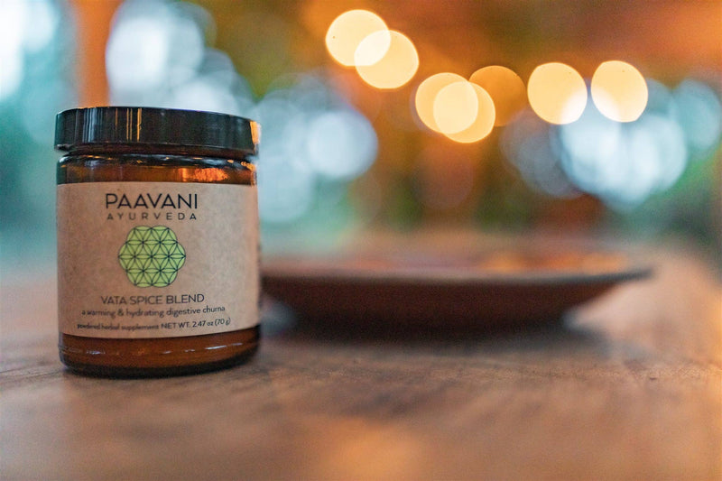Ein Glas "PAAVANI AYURVEDA VATA SPICE BLEND" (2.47 oz / 70g) steht auf einer Oberfläche, mit unscharfen, warm leuchtenden Lichtern im Hintergrund, die eine gemütliche Atmosphäre schaffen.