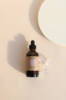 Eine braune Glaspipettenflasche mit Blumenwasser steht neben einem kleinen Glasbecher auf einem hellbeigen Hintergrund mit einem weißen Kreis in der Ecke.