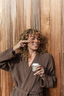  Eine lächelnde Frau mit lockigem, braunem Haar und einem braunen Bademantel trägt mit ihrem Finger eine dunkle Gesichtsmaske auf ihre Wange auf, vor einem Holzhintergrund.