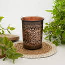 Dunkler Kupferbecher mit einem komplizierten geätzten Muster, der auf einem gewebten Untersetzer liegt und von grünen Pflanzen umgeben ist.