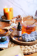 Eine kleine Flasche „Blue Lotus Tincture“ steht auf einem Holztablett mit einem großen, rauen blauen Kristall und einem klaren Glasbecher mit bernsteinfarbenem Tee, der blaue Lotusblüten enthält. Hohe brennende Kerzen sind im Hintergrund sichtbar.