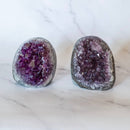Zwei grob herzförmige, geschnittene Amethyst-Geodenstücke stehen aufrecht auf einer weißen Marmoroberfläche. Beide zeigen im Inneren funkelnde, tief violette Kristalle, die von einem kontrastierenden grau-weißen, unpolierten Rand umgeben sind.