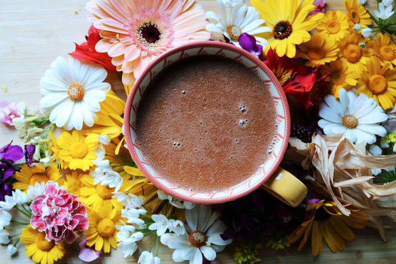  Eine Draufsicht auf eine Tasse mit schaumiger heißer Schokolade. Die Tasse ist in ein Bett aus bunten Blumen, darunter gelbe, weiße und rosa Gänseblümchen, auf einer hellen Holzoberfläche gebettet.