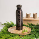Dunkle Kupfer-Wasserflasche, geätzt mit den sieben Chakra-Symbolen, präsentiert auf einem Holzbrett, umgeben von immergrünem Laub.