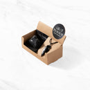 Eine offene, schräge Ansicht einer braunen Papp-Geschenkbox für das Set „Gift of Protection“ Schwarzer Obsidian, die einen Rohstein und einen passenden Pfeilspitzen-Anhänger an einer schwarzen Kordel enthält.