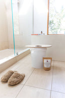 Ein weißer Eimer mit einem Holzsitz zum Dampfbad und ein Beutel mit Kräutermischung stehen auf einem gefliesten Badezimmerboden neben einem Paar flauschiger Hausschuhe.