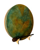 Eine runde Handtrommel (Rahmen- oder Schamanentrommel) mit einem bemalten Fell in gesprenkelten Petrol-, Grün- und Gold-/Orangetönen, schräg auf einem kleinen Holzständer ruhend, mit einem dunklen Holz-Schlägel davor platziert.