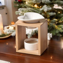 Eine Aromalampe aus Bambusholz mit einer herzförmigen Keramikschale, auf einem Holztisch vor einem funkelnden, leicht beschneiten Weihnachtsbaum.