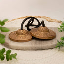 Ein Paar kleiner, verzierter Tingsha-Zimbeln aus Messing oder Bronze, verbunden durch eine schwarze Lederschnur, liegend auf einem Marmortablett und einer geflochtenen Matte.