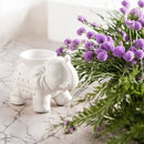 Weiße Elefanten-Duftlampe auf Marmorplatte neben einem Bündel lila Blumen.