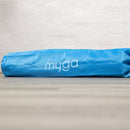Eine leuchtend blaue, zylindrische Yogamatten-Tasche mit dem weißen „myga“-Logo liegt horizontal auf einer Holzoberfläche vor einer strukturierten weißen Wand.