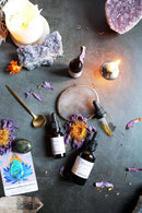 Eine Draufsicht zeigt eine mystische Anordnung auf einer dunklen, strukturierten Oberfläche. Zwei Flaschen "Blue Lotus Tinktur", ein goldener Löffel, ein polierter Labradoritstein und eine Orakelkarte mit einem blauen Lotusdesign sind prominent. Verschiedene Kristalle, darunter Amethystcluster und klarer Quarz, sind ebenfalls sichtbar, zusammen mit verstreuten getrockneten violetten Blütenblättern, orangen Safranfäden und brennenden Kerzen, die eine spirituelle Atmosphäre hervorrufen.