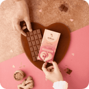 Zwei Hände halten eine Delikao "Love Tonic" Milchschokoladentafel mit Karamell- und Ingwerstückchen und deren Verpackung, platziert auf einem herzförmigen Tablett. Ingwerwurzel und -pulver sind unten links verteilt.