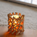 Ein rustikaler Kerzenhalter aus hellen, ineinandergreifenden Holzstäbchen, der auf einer Holzoberfläche vor einem weichen, weißen Teppich warm leuchtet.