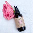 Eine braune Sprühflasche "PAAVANI AYURVEDA ROSE WATER soothing aromatherapy" (4 fl. oz.) steht neben einer großen, aufgeblühten pinken Rose auf einem weißen Tuch.