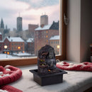 Ein beleuchteter Buddha-Zimmerbrunnen auf einem verschneiten Fensterbrett mit Blick auf eine verschneite Stadt.