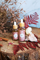 Zwei Flaschen "Friya Skincare Oil" stehen auf einem Holzscheit, umgeben von getrockneten Kräutern, Blumen und einem weißen Rosenquarz-Kristall.