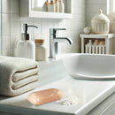 Ein rosa Himalaya-Salzseifenstück liegt auf einer weißen Waschtischplatte mit einem Häufchen Salz, einem gestapelten Handtuch und einem Seifenspender neben dem Waschbecken.