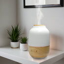 Aromatherapie-Diffusor aus Holz und weißer Keramik, der Dampf abgibt, neben zwei kleinen Pflanzen.
