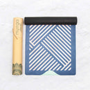 Eine teilweise ausgerollte Myga Yogamatte mit einem blau-hellrosa/weißen geometrischen Streifenmuster, die neben ihrer hohen, gebrandeten braunen Kartontragrolle liegt.