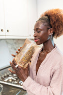 Eine Frau in einem rosafarbenen Bademantel lächelt, während sie an einem durchsichtigen Beutel mit getrockneten Kräutern und Blütenblättern in einer hellen Küche riecht.