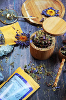 Eine offene Holzdose, gefüllt mit losem Kräutertee, steht auf einem dunklen Holztisch. Sie ist umgeben von Teepackungen, verstreuten Kräutern, getrockneten Blumen und Kristallen.