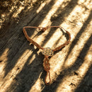 Ein handgefertigtes, braunes Makramee-Armband mit einem goldenen, filigranen Blumen des Lebens-Anhänger liegt auf einer Holzoberfläche mit Sonnenlicht und Schatten.