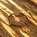 
Ein Armband mit einem bronzefarbenen Lotusblumen-Anhänger, geflochten in schwarzer Schnur, liegt auf einem Holzuntergrund.