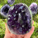 Eine Hand hält eine große, polierte, eiförmige Amethyst-Geode oder -Cluster mit tief königs-violetten Kristallen und einem klaren/weißen Quarzrand. Mehrere andere kleinere, dunkle Amethyst-Geodenfragmente sind auf dem hellgrünen Rasen im Hintergrund sichtbar.