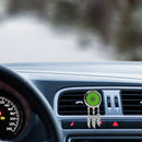 Ein silberfarbener Aromadiffusor im Traumfänger-Stil mit floralem Ausschnitt über einem leuchtend grünen Filzpad und drei silbernen Federanhängern, befestigt an einer Auto-Lüftungsdüse während der Fahrt.