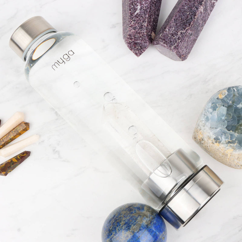 Eine schräg liegende "myga" Wasserflasche mit einem klaren Kristall, umgeben von verschiedenen Kristallen und Mineralien auf einer weißen Marmorplatte.