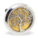 Eine Nahaufnahme eines runden, silberfarbenen Aromadiffusors für die Autolüftung, der ein kunstvolles ausgeschnittenes Lebensbaum-Design über einem hellgelben Filzpad zeigt.