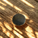 Ein runder, dunkler Anhänger aus poliertem Stein, der auf einer Holzoberfläche liegt, während Sonnenlicht ein Muster von Schatten erzeugt.