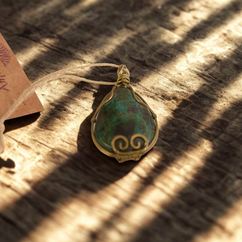 Ein birnenförmiger Anhänger mit einem türkisfarbenen Stein, der von goldfarbenem Draht mit Spiralverzierungen umwickelt ist, liegt auf einer Holzoberfläche mit starken Schattenstreifen. Links oben ist ein Teil eines braunen Etiketts mit Schnur sichtbar.