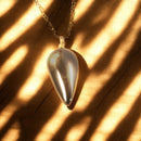 Ein tropfenförmiger silberner Anhänger, der an einer Kette hängt, beleuchtet von sanftem Licht, das seine glatte Oberfläche hervorhebt und Schattenmuster im Hintergrund erzeugt.