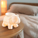 Beleuchtete weiße Keramik-Duftlampe in Elefantenform auf einem Nachttisch neben einem Bett.
