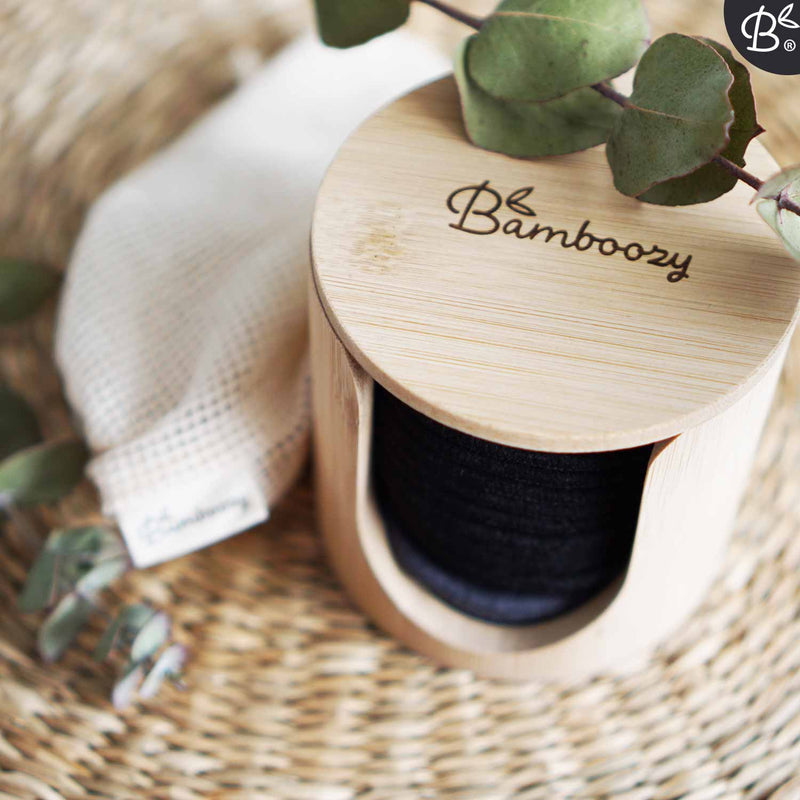 Nahaufnahme von Bamboozy wiederverwendbaren Make-up-Entfernerpads in einem Bambushalter und einem Netzwaschbeutel auf einer geflochtenen Oberfläche, mit einem grünen Zweig.