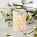 Ein weißer, leuchtender elektrischer Wachswärmer mit einem kunstvollen, wirbelnden floralen Ausschnittmuster, der vor einem weichen, weißen Hintergrund mit unscharfen weißen Blüten und grünen Stielen steht.