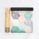 Eine teilweise ausgerollte Myga Yogamatte mit einem Mosaikmuster aus pastellfarbenen Sechsecken mit Marmor- und strukturierten Oberflächen, die neben ihrer braunen Kartontragrolle liegt.