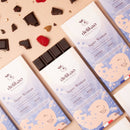 Ein Delikao "Super Woman" Schokoladenriegel und seine Verpackung liegen auf einer rosa Oberfläche, umgeben von verstreuten Mandeln, Himbeeren, blauem Borage und Schokoladentropfen. Eine Hand liegt im Vordergrund.