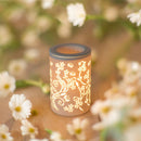 Eine beleuchtete, keramische Duftlampe/Wärmediffusor mit einem ausgeschnittenen Blumenmuster steht auf einer Holzoberfläche, umgeben von unscharfen weißen Blüten.