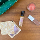 Flat Lay von Schönheitsprodukten, darunter Paavani Ayurveda Women's Moon Oil, ein Taschenspiegel und Lippenfarbe auf einem Holztisch.