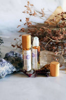 Zwei Roll-on-Flaschen mit Chakra-Öl und Holzdeckeln, umgeben von getrockneten bräunlichen Blumen, einem Bund getrocknetem Lavendel, einem großen blauen Fluoritkristall und einem Quarzkristall auf weißem Hintergrund.