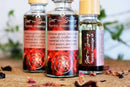 Drei Tropfflaschen "Hot n Spicy Tantric Massage Oil" sind auf einer Holzoberfläche präsentiert, wobei die mittlere Flasche scharf im Fokus liegt. Die Etiketten haben einen roten Hintergrund mit weißem Text, der Inhaltsstoffe wie Hibiskusblüten, Rizinusöl und ätherische Öle detailliert beschreibt. Getrocknete Blütenblätter sind um die Flaschen verstreut und tragen zur natürlichen Ästhetik bei.