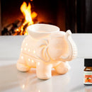 Beleuchtete weiße Keramik-Duftlampe in Elefantenform neben einem Fläschchen Orangenöl vor einem Kamin.